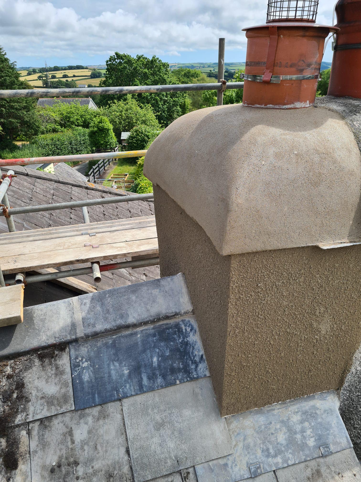 A close-up of a newly rendered chimney on a tiled roof, with scaffolding and countryside views, including green trees and fields, in the background under a partly cloudy sky.