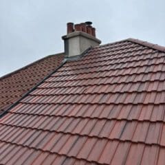 A sloped roof covered with red tiles is shown against a cloudy sky. A chimney with several pots is visible near the top of the roof.