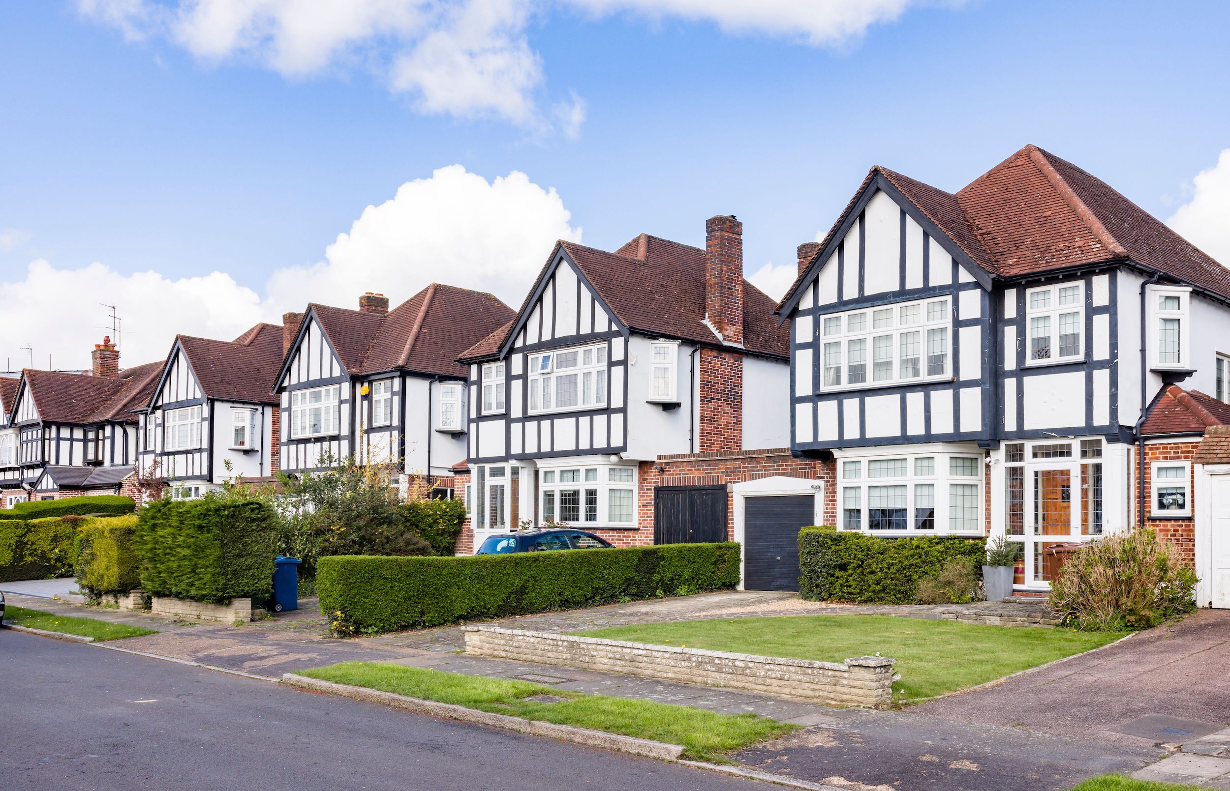 A row of traditional two-story Tudor-style houses with black and white timber framing. Each has a neat front lawn and driveway. The sky is clear with a few clouds.