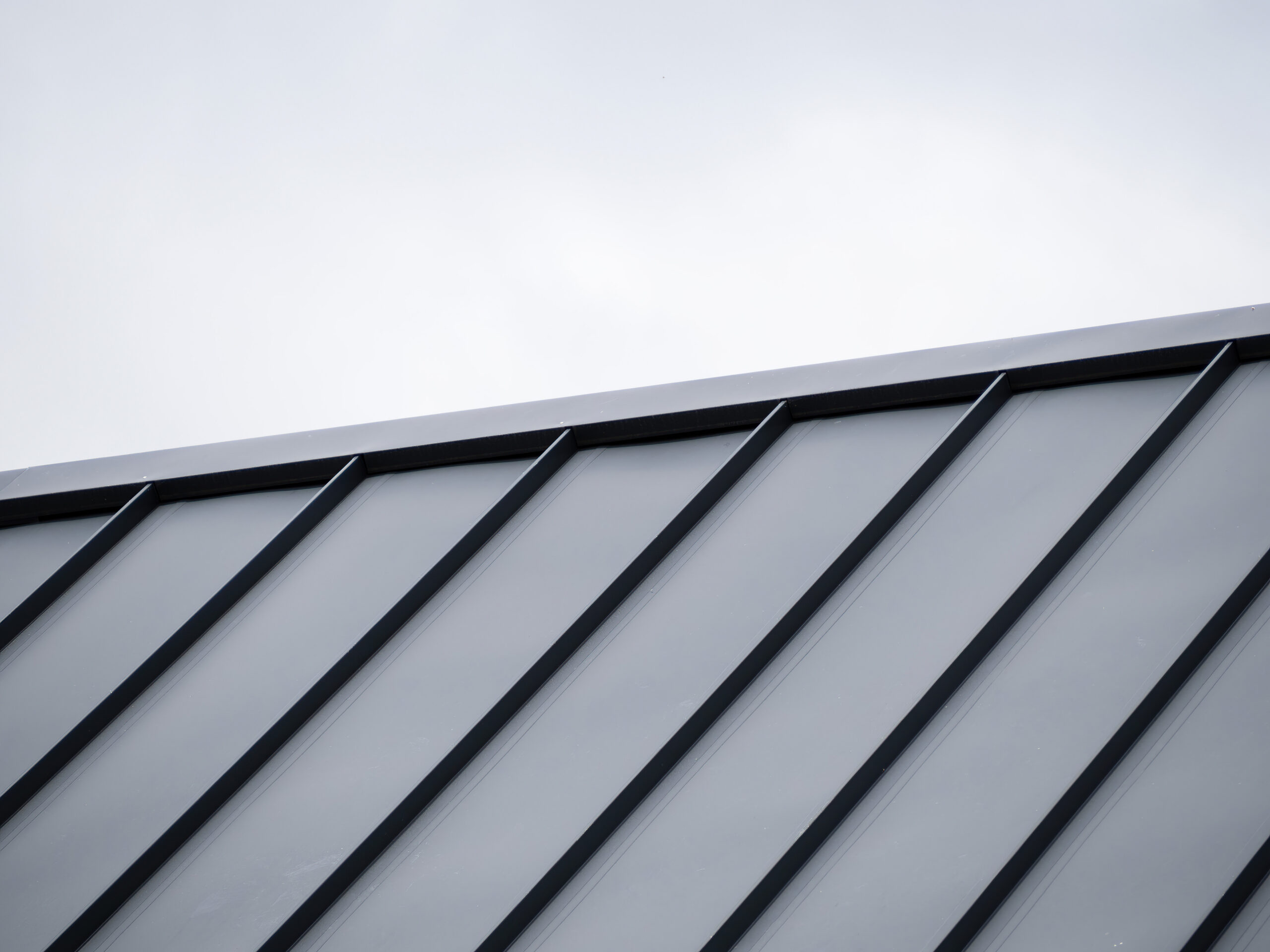 A close-up of a modern metal roof with angled black seams and panels, set against a cloudy, light gray sky.