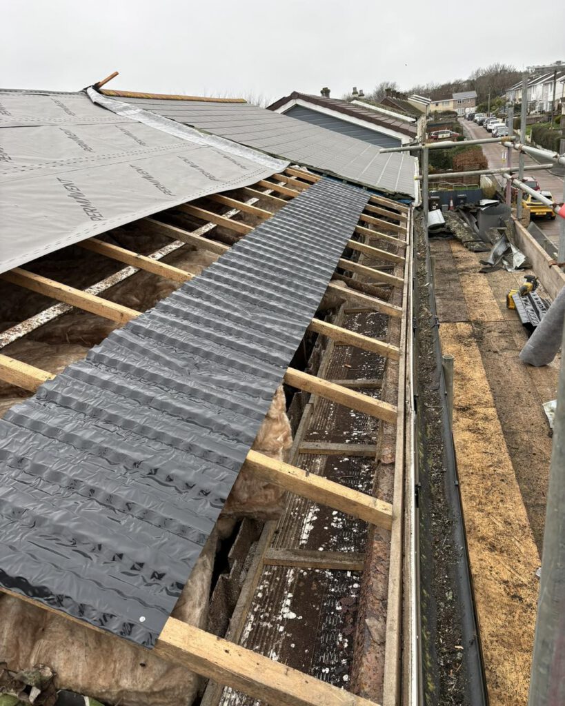 A partially exposed roof under construction, with insulation and wooden beams visible. A strip of protective waterproof material runs along the center, and construction materials are scattered nearby. Houses and a road are seen in the background.