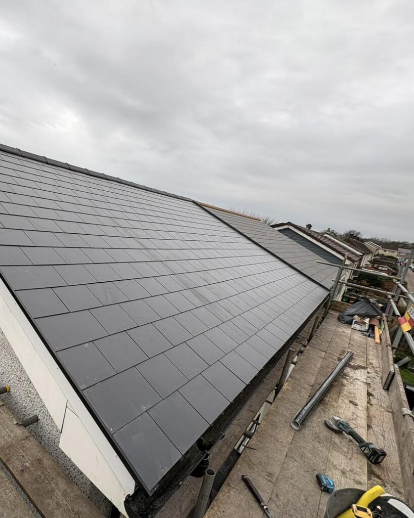 A newly installed slate tile roof on a residential building under cloudy skies, with construction tools and materials scattered on wooden scaffolding along the roof edge.