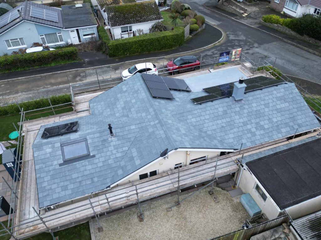 Aerial view of a house with a gray slate roof featuring solar panels, skylights, a chimney, and surrounded by scaffolding. Neighboring houses and a street with parked cars are also visible.