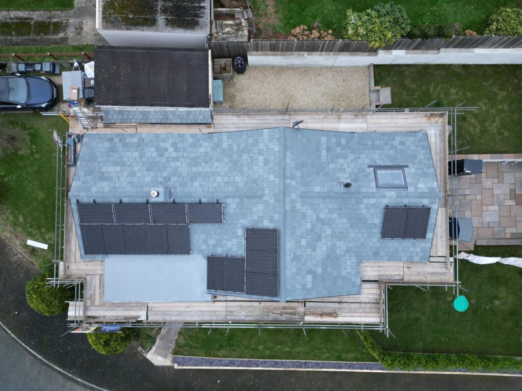 Aerial view of a house roof with gray shingles, multiple solar panels, a skylight, and surrounding scaffolding. The property has a driveway, garden, and is bordered by a fence and neighboring structures.