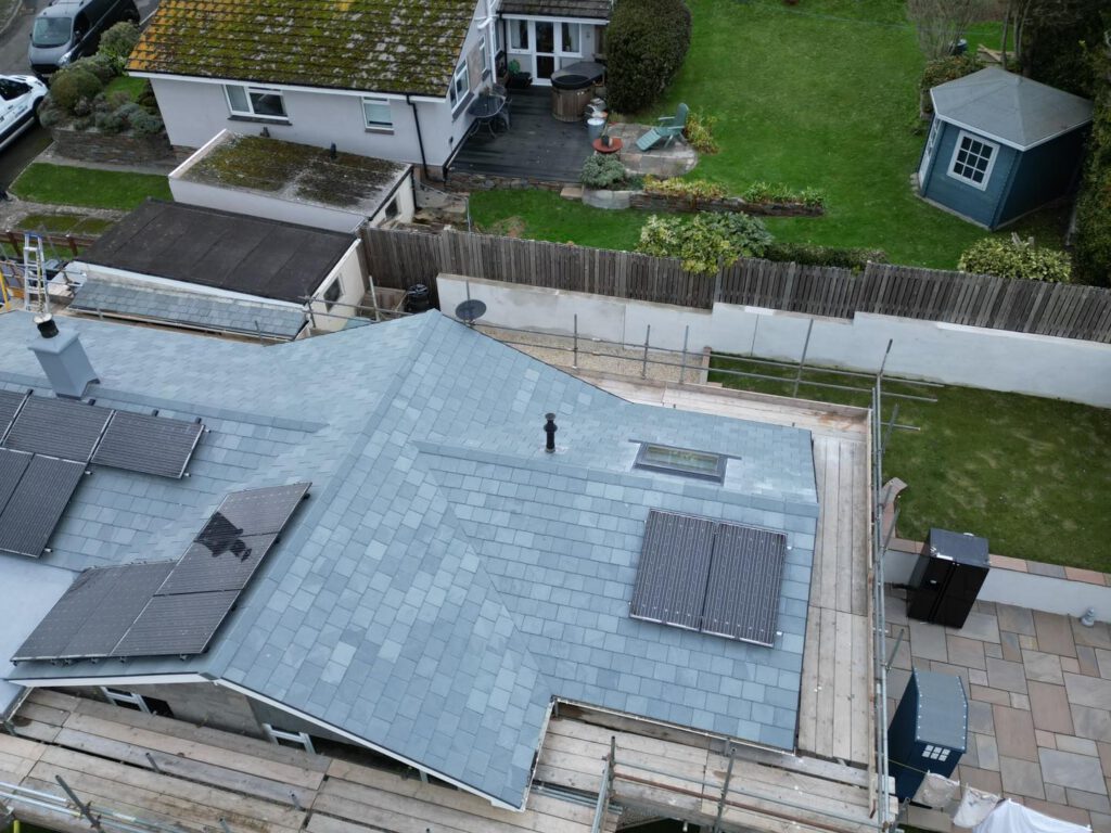 Aerial view of a house with a grey slate roof, solar panels, and scaffolding around it. The backyard has green grass, a patio, a shed, and a house with moss on its roof in the background.