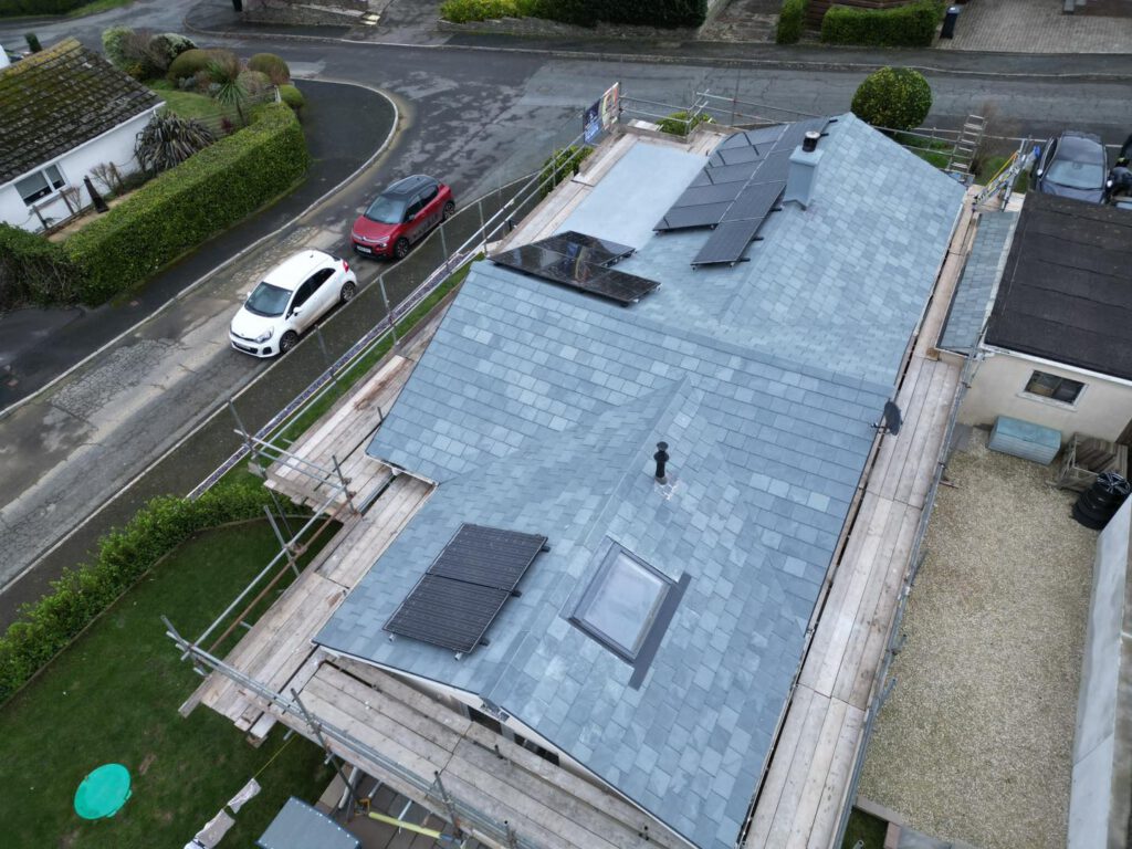 Aerial view of a house with a gray slate roof, solar panels, skylights, and scaffolding surrounding the building. Cars are parked on the nearby road and the neighborhood is visible in the background.
