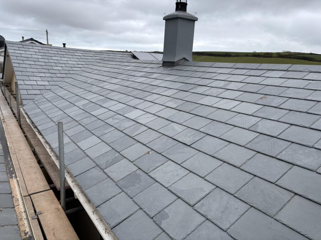 A newly installed slate roof with neatly arranged grey tiles, a central chimney, and a distant view of green fields under a cloudy sky. Scaffolding is visible along the roof edge.