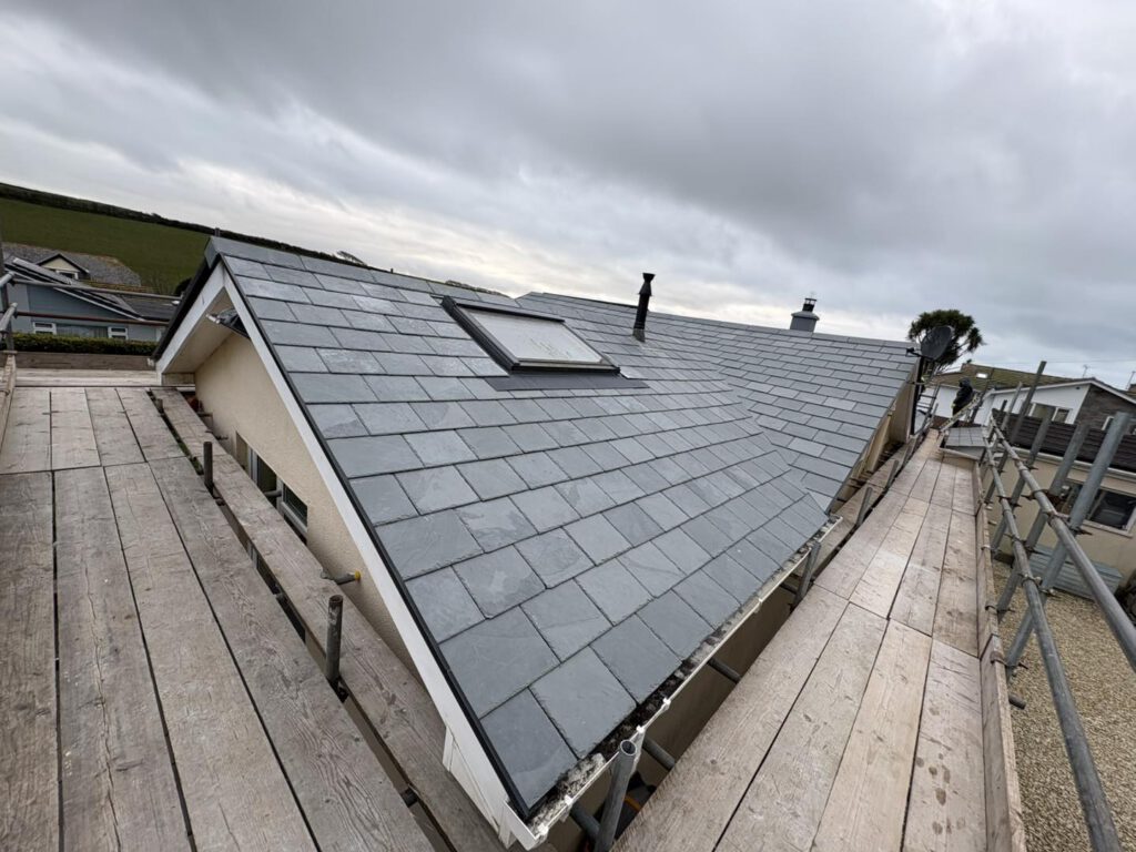 A newly installed slate roof on a house, viewed from scaffolding under a cloudy sky. The roof features a skylight, a chimney, and is surrounded by rural landscape and nearby buildings.