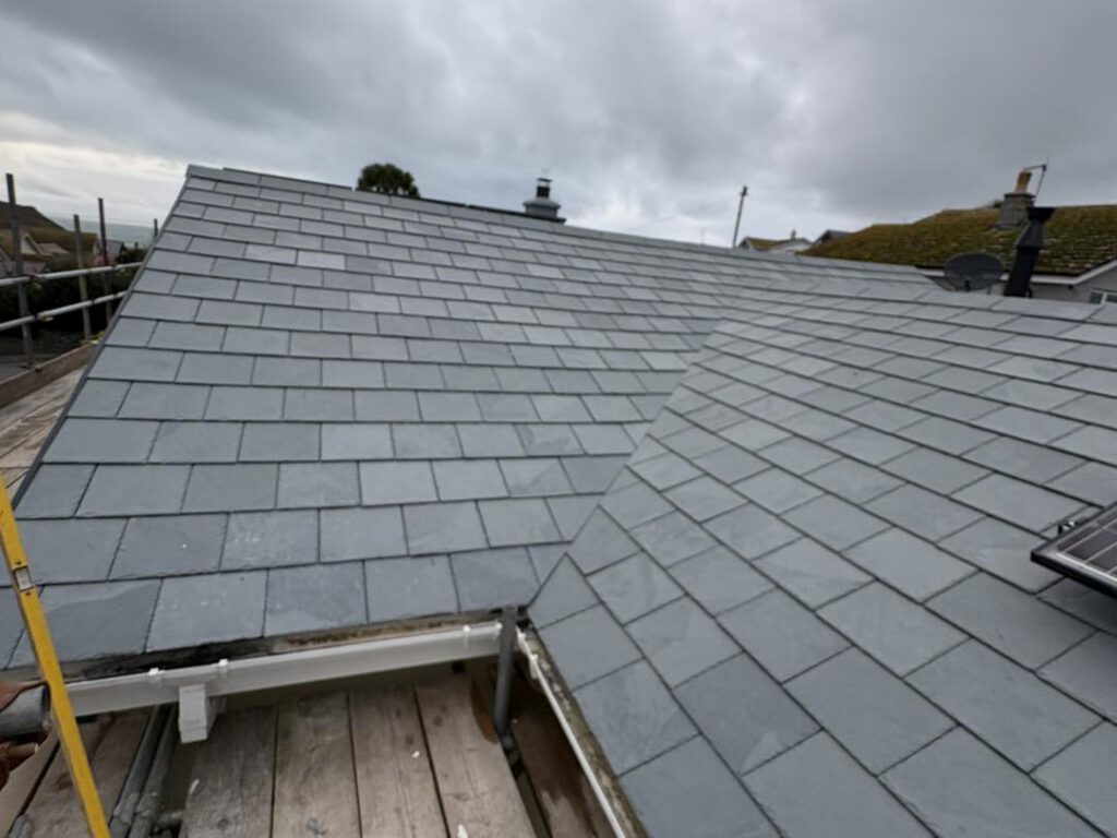 A close-up view of a grey slate-tiled roof under a cloudy sky, with scaffolding and part of a wooden platform visible in the foreground.